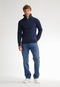 Marine gebreide trui met een ritssluiting kraag en ribbelzoom. Draagt bij blauwe jeans en witte sneakers, met een lichte gestructureerde textuur.