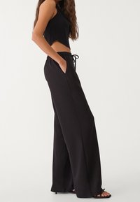 Femme portant un pantalon noir à jambes larges avec taille à cordon et un top court noir sans manches, debout avec une main dans la poche.