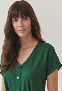 Groene V-hals knoopshirt met korte opgerolde mouwen, gemaakt van zachte stof. Kenmerkt zich door bijpassende knopen aan de voorkant.