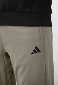 Jasnooliwkowe spodnie sportowe z teksturowanym wykończeniem, z czarnym logo Adidasa w pobliżu dołu i czarną elastyczną talią.