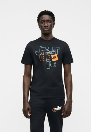 Muž nosi čierne tričko Nike s modro-oranžovým textom "Just do it" a čierne nohavice s oranžovým logom Nike, stojaci pred bielym pozadím.
