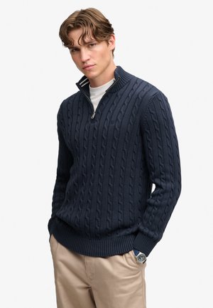 Jeune homme portant un pull bleu marine en maille torsadée à demi-fermeture éclair, pantalon beige, sous-vêtement blanc et une montre-bracelet, les mains dans les poches.