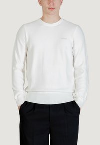 Sweat-shirt blanc en coton à col rond avec poignets et ourlet côtelés, ornée d'un logo brodé discret sur la poitrine. Associé à un pantalon foncé.