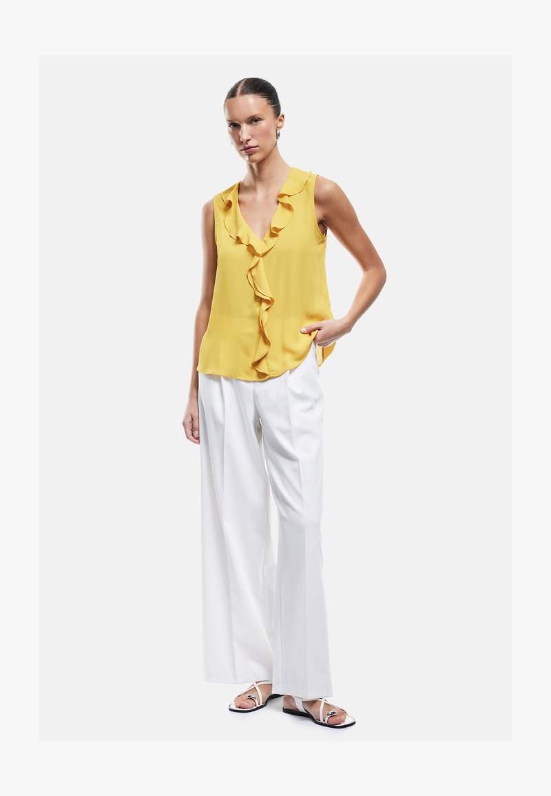 Femme portant un chemisier jaune sans manches à volants, un pantalon blanc à jambes larges et des sandales blanches, debout avec une main dans la poche.