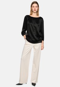 Woman standing with hands in pockets wearing black satin blouse, white wide-leg trousers, and black pointed-toe heels on white background.