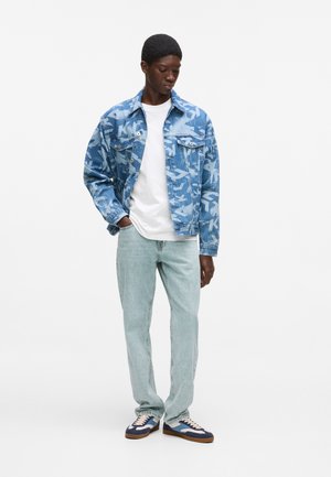 Jeune homme portant une veste en denim bleue à motifs, un t-shirt blanc, un jean bleu clair et des baskets bleues et blanches, debout devant un fond blanc.