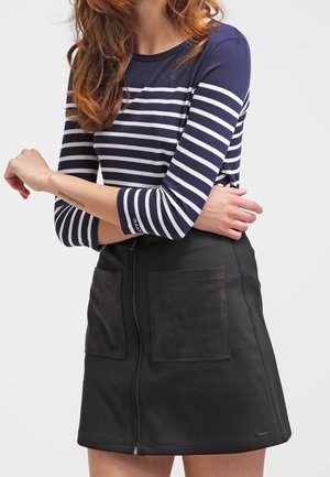 Mujer con camiseta de manga larga a rayas azul marino y blanca y falda negra con cremallera frontal y grandes bolsillos parche, con los brazos cruzados.