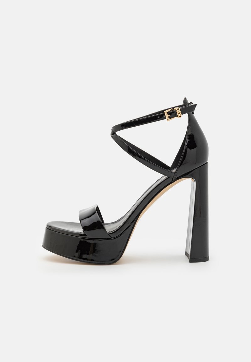 Sandale à talons hauts en cuir verni noir avec semelle plateforme, bride à la cheville et boucle dorée sur fond clair.