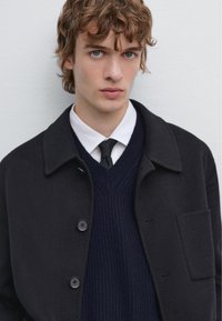 Sort uldblandingsjakke med krave, der har en knaplukning og en påsyet lomme, lagdelt over en marineblå v-hals trøje og en hvid skjorte.