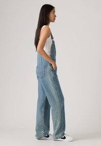 Τζιν ολόσωμη φόρμα σε ανοιχτό μπλε, με ρυθμιζόμενους ιμάντες, μπροστινή τσέπη και άνετη εφαρμογή. Συνδυάζεται με λευκό αμάνικο τοπ και αθλητικά παπούτσια.