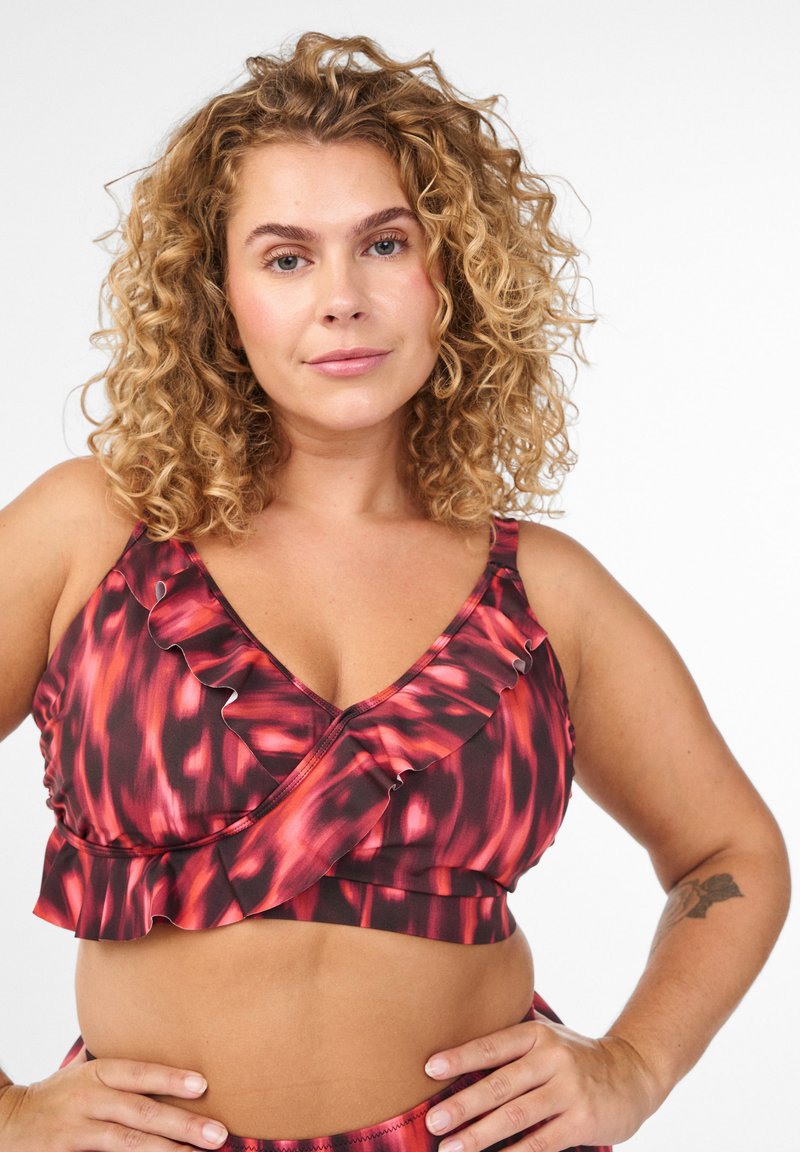 Rød og sort mønstret bralette med flæsedetaljer, udstyret med V-udskæring og blødt stof. Modellen har krøllet hår.