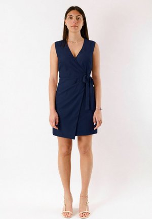 Donna con capelli lunghi e scuri in piedi di fronte, indossa un vestito blu notte senza maniche con un nodo laterale e sandali beige con tacco alto.