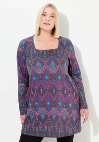 Langarm tuniek gemaakt van rekbaar materiaal, met een vierkante halslijn en een ingewikkeld geometrisch patroon in paars, rood en blauw op een donkere achtergrond.