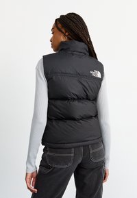 Vesta neagră tip puffer cu guler înalt, având panouri cusute și un logo alb pe spate. Purtați-o peste un tricou cu mâneci lungi de culoare gri deschis.