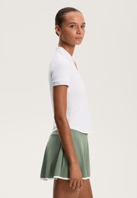Biała koszulka polo z krótkim rękawem, w połączeniu z zieloną spódnicą w kształcie litery A z białym wykończeniem. Gładka tekstura, casualowy sportowy design.