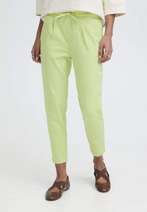 Personne portant un pantalon vert clair longueur cheville avec une taille à cordon et des ballerines marron à lanières croisées sur fond blanc.