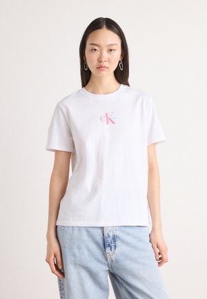 Bílý bavlněný T-shirt s krátkým rukávem a kulatým výstřihem, na přední straně s barevným logem CK. V kombinaci s světle modrými džíny.