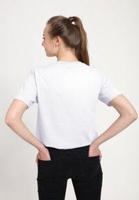 Bílé bavlněné tričko s krátkými rukávy, kulatým výstřihem a volným střihem. Nošeno s černými džínami, ruce v zadních kapsách.