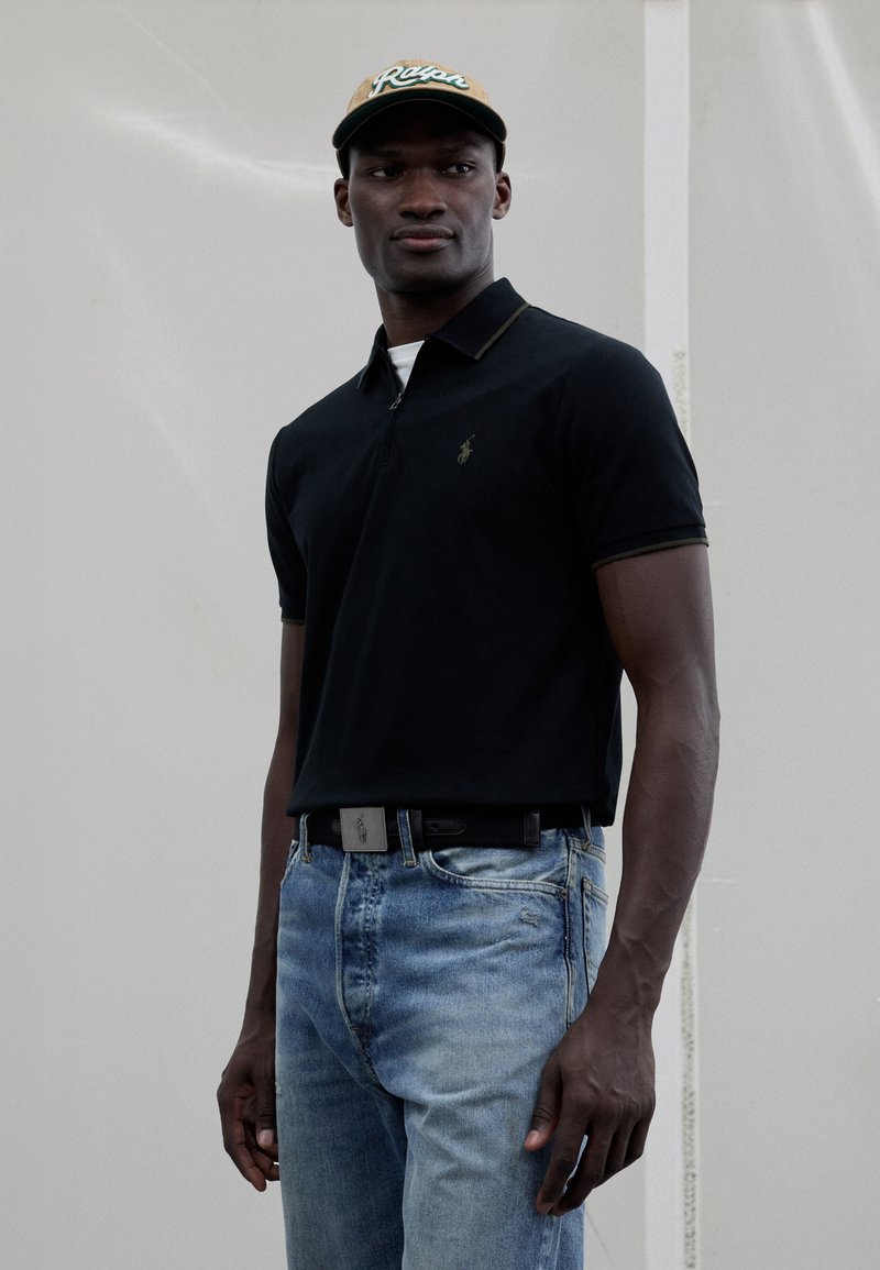 Svart polotrøye med glidelåshals, grønne detaljer, kombinert med lyseblå denimjeans. En beige baseballcap med grønn tekst fullfører antrekket.