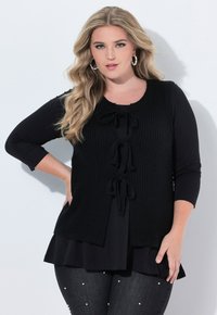 Svart stickad väst med knytning fram, lager på lager över en svart topp. Långärmad och strukturerad kropp. Jeansen har pärlaccenter.