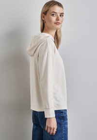 Helle beige Kapuzenjacke mit lockerer Passform, weicher Textur und Kängurutasche, kombiniert mit dunklen Jeans. Einfaches Design, keine Muster oder Akzente.