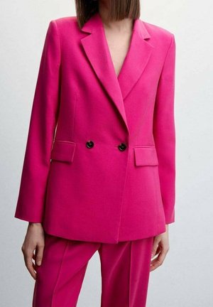 Kvinde iført en skarpt pink skræddersyet blazer med sorte knapper og matchende bukser mod en ensfarvet lys baggrund.
