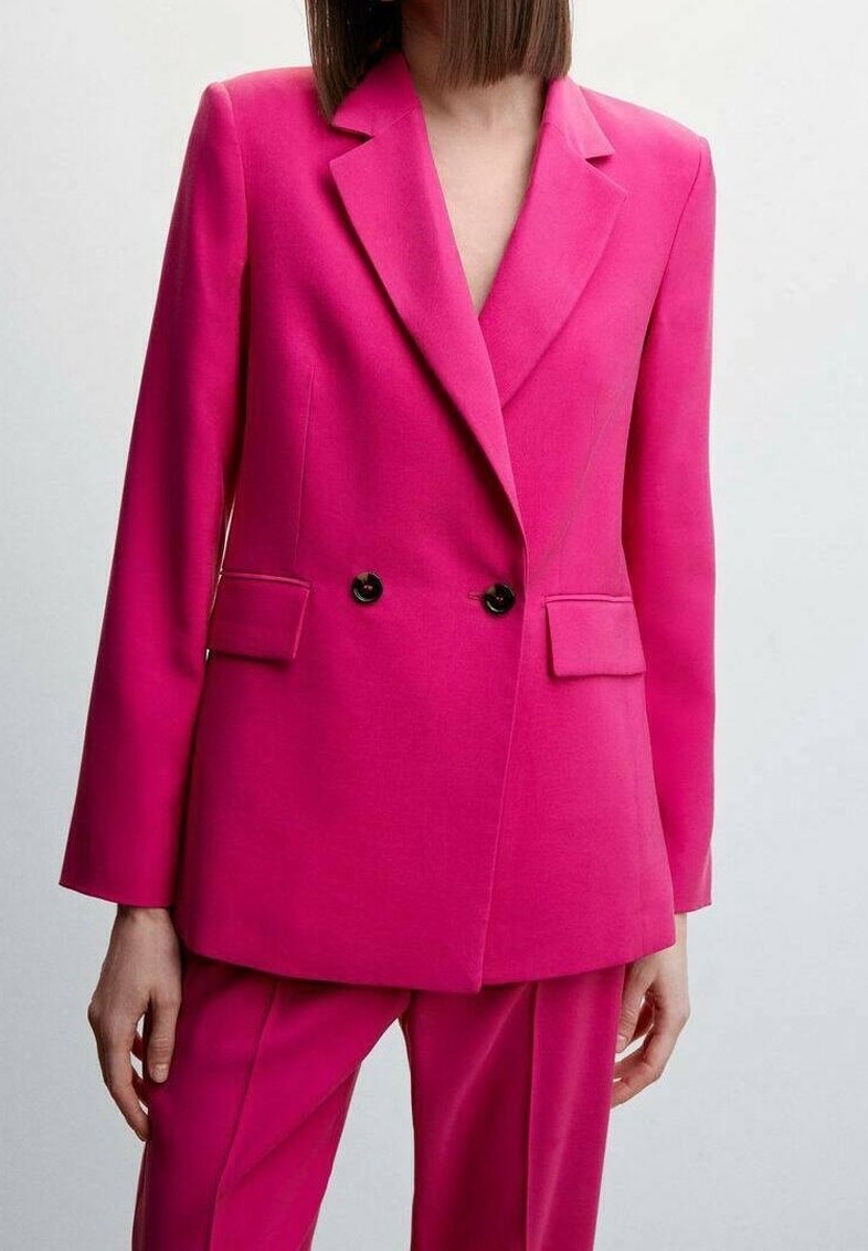 Femme portant un blazer rose vif cintré avec des boutons noirs et un pantalon assorti sur un fond clair uni.