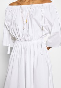 Femme portant une robe blanche décolletée avec taille froncée et liens à pampille, accessoirisée d'un collier en chaîne dorée avec pendentif.