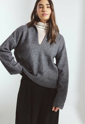 Mladá žena s dlhými hnedými vlasmi nosiaca zlaté kruhové náušnice, biely rolák, sivý sveter s výstrihom do V a čiernu sukňu na neutrálnom pozadí.