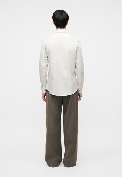 Άτομο με κοντά μαύρα μαλλιά που φορά λευκό μακρυμάνικο πουκάμισο και χαλαρό καφέ παντελόνι, στέκεται με την πλάτη προς τα εμπρός σε απλό φόντο.