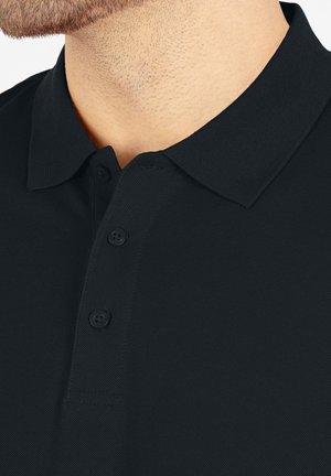 Zwarte poloshirt van een textuurstof, met een drieknoopsluiting en een klassieke kraag. Close-up weergave toont de stikseldetails en knopen.