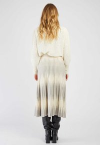 Kobieta z długimi falowanymi blond włosami, ubrana w sweter w kremowy wzór, plisowaną spódnicę w odcieniach beżu i szarości oraz czarne botki na obcasie, stoi na tle białym.