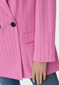 Blazer rosa a righe con un design doppiopetto, bottoni neri e una tasca frontale. Il tessuto appare liscio con una sartoria strutturata.