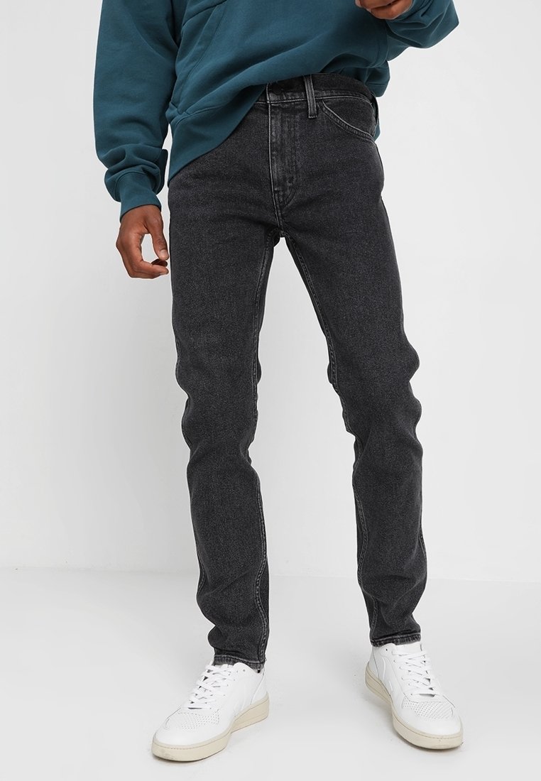 Homme portant un jean slim noir délavé, des baskets blanches et un sweat bleu-vert sur un fond clair uni.