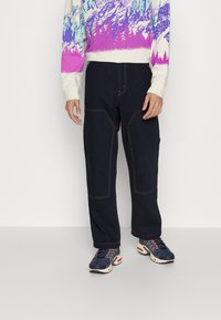 Pantalones de mezclilla azul oscuro con costuras visibles, forma cuadrada y bolsillos delanteros de parche, combinados con un suéter estampado multicolor y zapatillas.