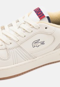 Baskets en cuir blanc cassé avec lacets blancs, doublure intérieure beige, semelle en gomme et logo crocodile sur le côté, étiquette rouge et marine sur la languette.