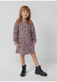 Ein junges Mädchen trägt ein langärmliges lavendelfarbenes Blumenkleid und schwarze, lacklederne Schnürstiefel und steht vor einem schlichten hellen Hintergrund.