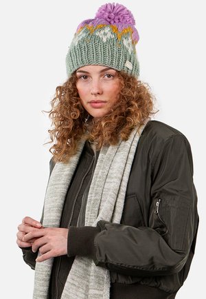 Bonnet tricoté vert avec un pompon violet, comportant des motifs multicolores. Associé à une veste bomber sombre et une écharpe grise claire.