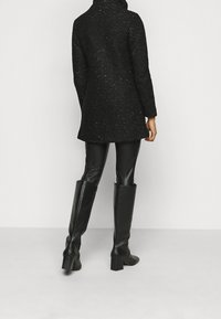 Svart texturerad kappa med hög krage, lös passform och långa ärmar, kombinerad med åtsittande svarta läderbyxor och knähöga klackstövlar.