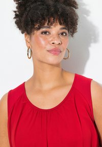 Top rosso senza maniche con scollo rotondo e dettagli arricciati. Il modello ha i capelli ricci e indossa grandi orecchini a cerchio dorati su uno sfondo bianco.