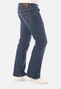 Donkerblauwe denim jeans met een uitlopende pijp, met zichtbare stiksels en een bruine merklabel aan de achterkant van de tailleband.