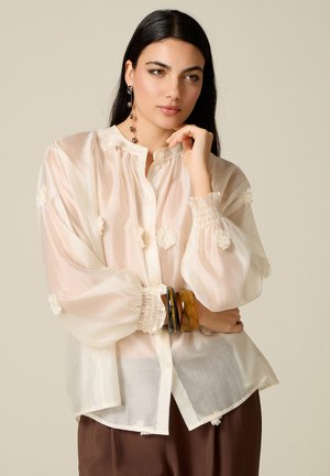Donna con capelli lunghi e scuri indossa una camicetta bianca trasparente con dettagli floreali, pantaloni marroni, orecchini pendenti e braccialetti spessi, posando pensierosa.