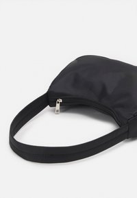 Czarna torba z nylonu o zakrzywionym kształcie, z zapięciem na zamek błyskawiczny i płaskim, szerokim paskiem. Gładka tekstura z minimalnymi detalami.