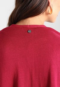Suéter burdeos con una textura de punto fino, con un escote redondo y un pequeño botón de logo negro en la parte posterior del cuello.