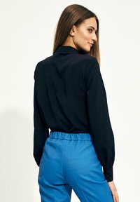 Czarna koszula z długim rękawem i kołnierzykiem, zestawiona z niebieskimi spodniami elastycznymi w pasie. Gładka tekstura tkaniny, minimalne detale i dopasowany krój.