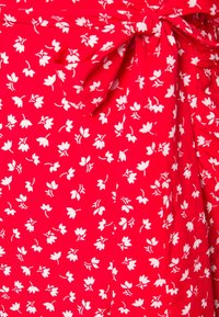 Robe en tissu rouge avec un motif floral blanc et un détail noué à la taille. Matière légère avec une texture fluide.