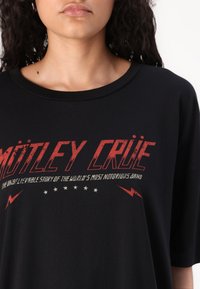 Must t-särk, millel on punane ja valge graafiline tekst "MÖTLEY CRÜE", välgu aktsentidega ja pehme puuvilla tekstuuriga.