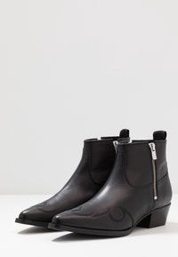 Botas de tobillo de cuero negro con punta afilada, cuentan con costuras decorativas y cremalleras laterales con herrajes plateados. Diseño de tacón bajo cuadrado.