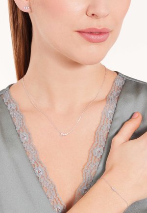 Bijoux en argent comprenant un collier avec une chaîne délicate agrémenté de trois petites gemmes et un bracelet assorti. Tissu gris avec des bordures en dentelle en arrière-plan.