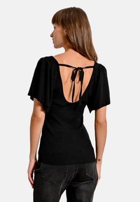 Top nero a coste con maniche a gufo e schiena scoperta, con chiusura a fiocco. Il tessuto ha una texture liscia e un design aderente.